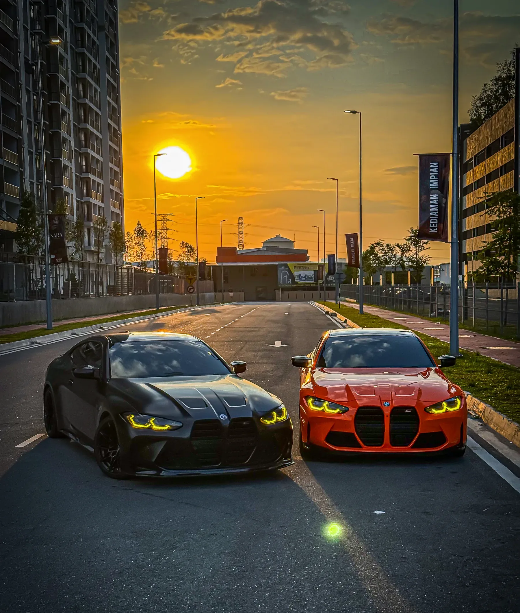Two BMW M4s, one black and one orange, on the road at sunset